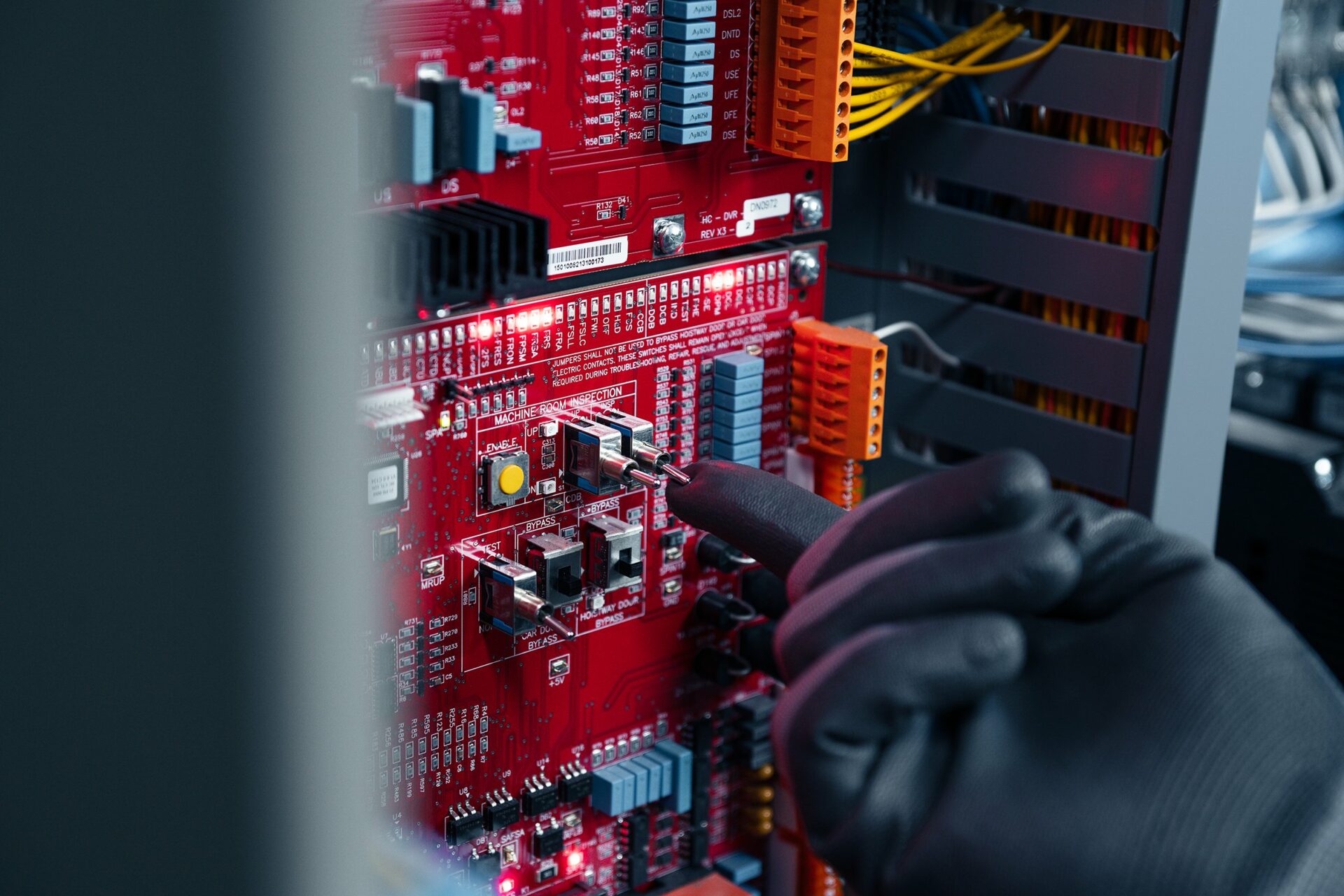 DSC07232-Edit copy A technician's hand adjusting components on a red circuit board within an elevator system.