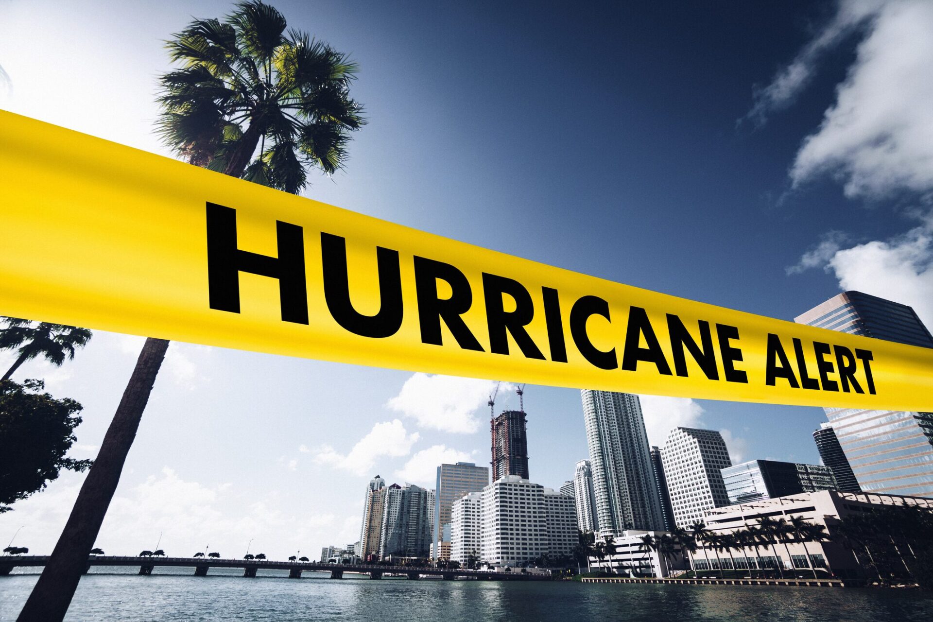 A yellow “Hurricane Alert” banner stretches across a city skyline with palm trees and high-rise buildings in the background.