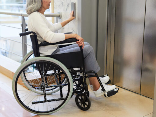 Older woman in a wheelchair positioned at an elevator entrance, reaching forward to press the call button.