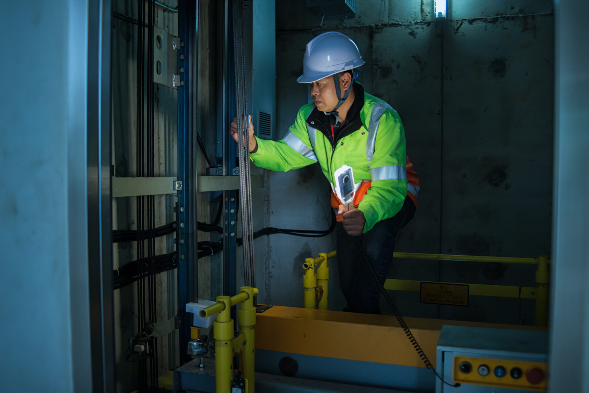 Elevator technician wearing a hard hat and high-visibility jacket inspecting cables and components inside an elevator shaft with a flashlight.