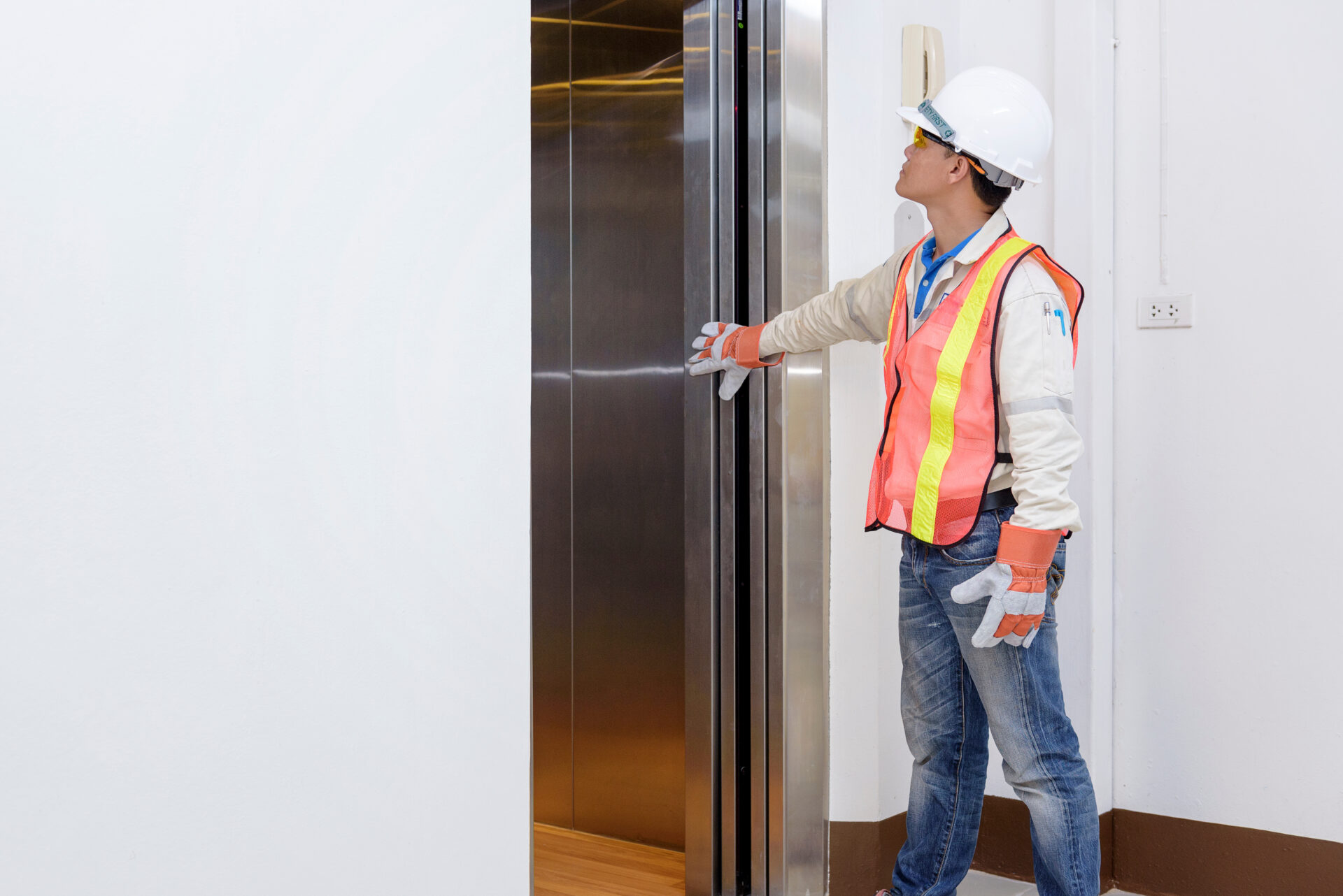 A maintenance worker in a safety vest and hard hat inspects an elevator door, ensuring it opens and closes properly.