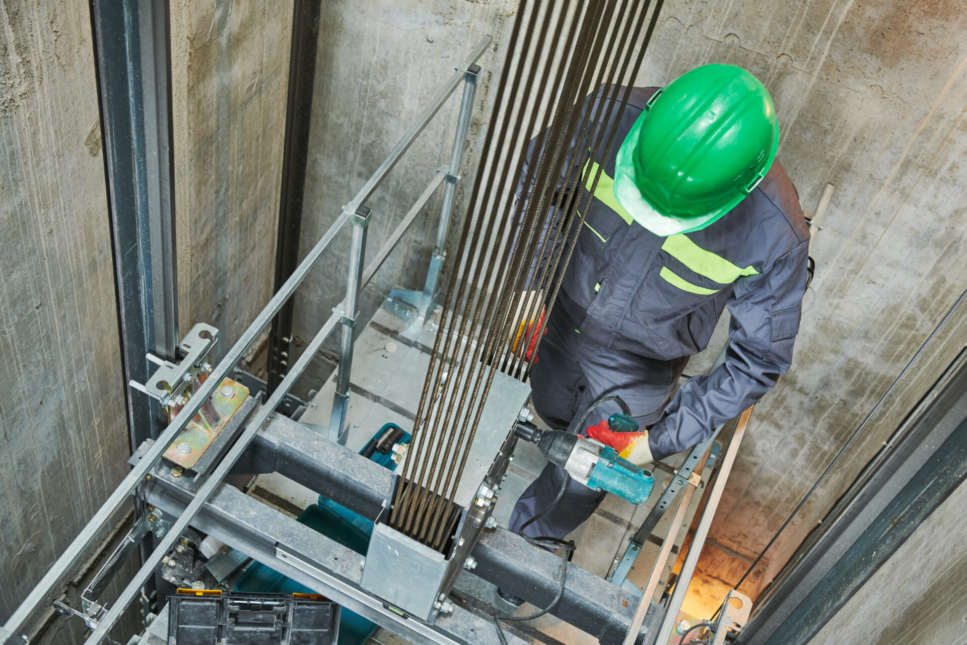 A technician wearing a green hard hat and safety uniform uses a power tool while servicing elevator cables inside the elevator shaft.